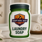 Soapy Mtn. Suds My Way Retreat Laundry Soap with luxury hotel-inspired fragrance, featuring a spa-like setting with white towels, candles, tropical greenery, and a coastal resort view.