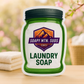 Soapy Mtn Suds April Fresh Downy Type Laundry Soap pouch on a wooden surface with folded towels, laundry basket, and spring blossoms in the background.