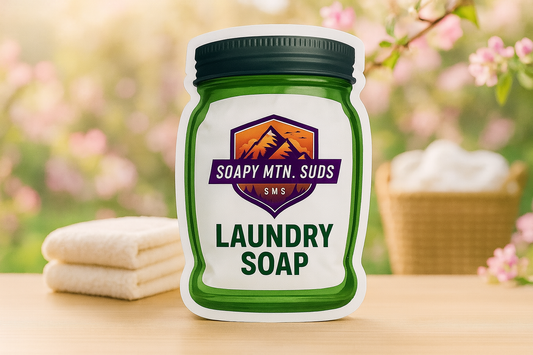 Soapy Mtn Suds April Fresh Downy Type Laundry Soap pouch on a wooden surface with folded towels, laundry basket, and spring blossoms in the background.