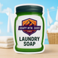 Soapy Mtn Suds Fresh AF Tide Type Laundry Soap pouch on a wooden surface with folded towels, a basket of cotton, and fresh laundry drying outdoors against a blue sky.