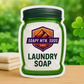Soapy Mtn Suds Fresh Fields Laundry Soap pouch on wooden surface with folded towels and green clovers in the background.