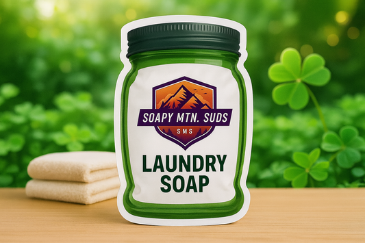 Soapy Mtn Suds Fresh Fields Laundry Soap pouch on wooden surface with folded towels and green clovers in the background.