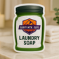 Soapy Mtn Suds Westin Type Laundry Soap in green mason jar bag packaging, hotel-inspired pH-neutral laundry powder displayed on marble counter with folded towels and spa-style candle.